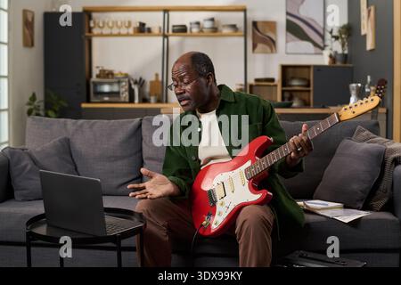 Middle aged Black man sitting on sofa playing electric guitar, while gesturing and speaking during online lesson using laptop in modern living room with shelves and abstract wall art Stock Photo