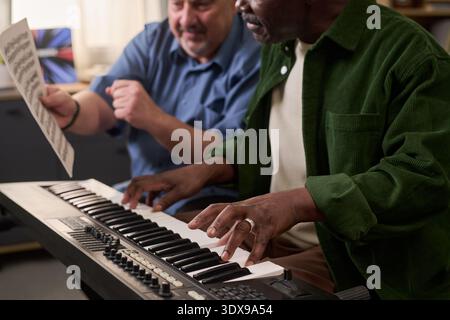 Middle aged Black man playing electronic keyboard while middle aged Caucasian man holding sheet music and gesturing, both focusing on musical performance in indoor setting Stock Photo