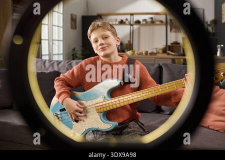 Portrait of Caucasian boy playing electric bass guitar while sitting on sofa, smiling and looking into camera, holding instrument with both hands, home interior visible in background Stock Photo