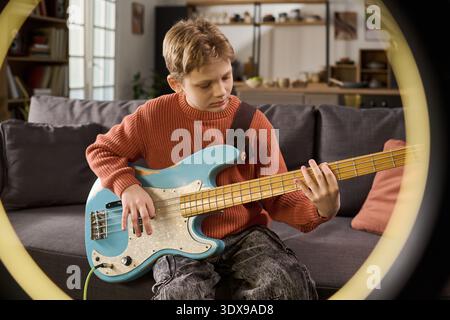 Caucasian boy playing electric bass guitar while sitting on sofa, focusing on finger placement and technique, demonstrating musical practice in home living room environment Stock Photo