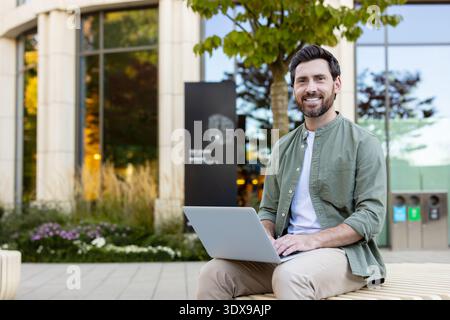 Happy young man smiling and looking at camera while working remotely on laptop, enjoying the casual work setting outdoors in a modern urban environment Stock Photo