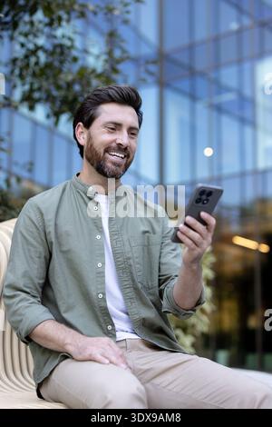 Happy bearded man smiling, engaging in a video call or browsing on his mobile phone while sitting casually on a contemporary bench outside a corporate glass building Stock Photo