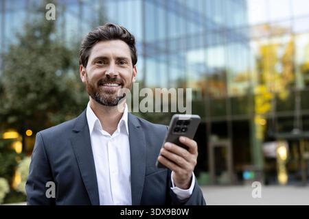 Executive man networking and communicating using a mobile phone, representing remote work, smart technology, and corporate communication in a professional outdoor setting Stock Photo