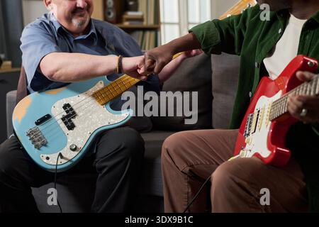 Middle aged Caucasian man holding electric bass guitar fist bumping middle aged Black man holding electric guitar sitting together on sofa, engaging in friendly interaction Stock Photo