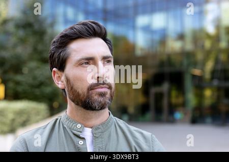 Bearded man with a serious, pensive expression looks away while standing outdoors against a modern office building, conveying confidence, determination and urban ambition Stock Photo
