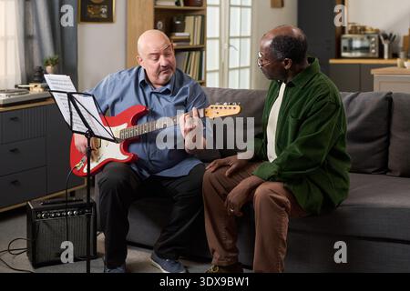 Middle aged Caucasian man playing electric guitar and reading sheet music, while sitting on couch next to senior Black man listening attentively in modern living room setting Stock Photo