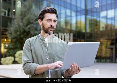 Man standing outdoors in front of a glass building, focused on typing on a laptop, representing remote work, flexible jobs, and mobile business through technology Stock Photo