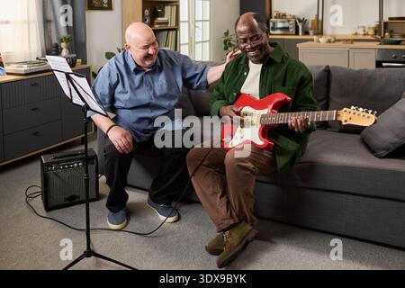 Middle aged Caucasian man smiling and encouraging middle aged Black man playing electric guitar on sofa, music stand and amplifier nearby, both enjoying music session indoors Stock Photo