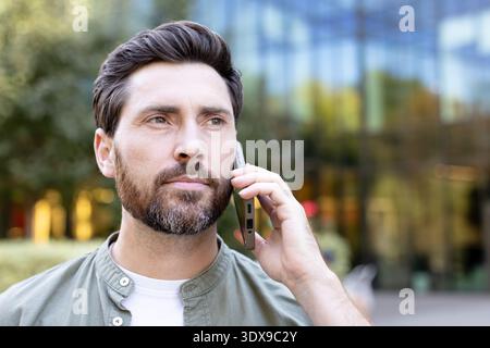 Man talking on a mobile phone with a serious facial expression, looking away while having a conversation in an urban environment with office buildings in the background Stock Photo