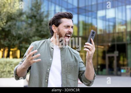 Bearded man shouting at his smartphone outdoors, expressing anger, frustration and stress from digital communication, bad news or a heated argument, intense and upset emotions Stock Photo