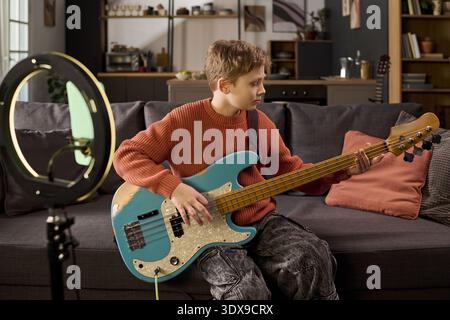 Caucasian boy playing electric bass guitar sitting on sofa, focusing on instrument, ring light positioned in foreground, home interior visible in background, casual setting Stock Photo