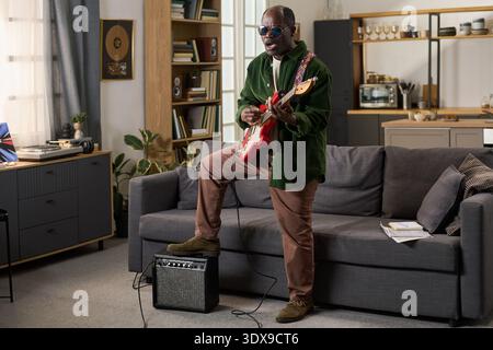 Middle aged Black man playing electric guitar while standing in living room, wearing sunglasses and singing, amplifier on floor, bookshelf and kitchen visible in background Stock Photo