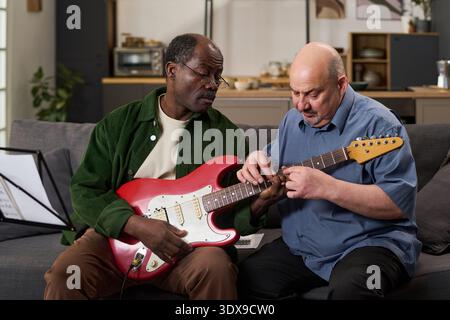 Middle aged Black man playing electric guitar while middle aged Caucasian man teaching him technique on sofa, both focused on instrument, music stand with sheet music visible nearby Stock Photo