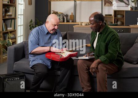 Middle aged Caucasian man demonstrating electric guitar technique to middle aged Black man sitting on sofa, Black man holding notebook and pen, amplifier on floor nearby Stock Photo