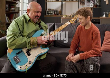 Caucasian middle aged man teaching Caucasian boy playing electric bass guitar while sitting together on sofa, man demonstrating finger placement, boy watching attentively, cozy living room background Stock Photo