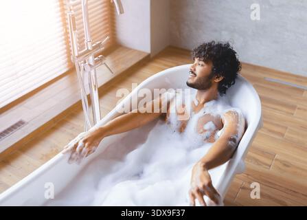 Man Relaxes in Bathtub With Bubbles and Sunlight in Modern Bathroom Stock Photo