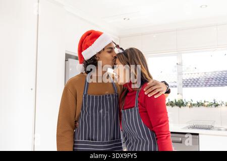 Diverse couple kissing and leaning in home kitchen, wearing striped aprons, Santa hat, smartwatch Stock Photo