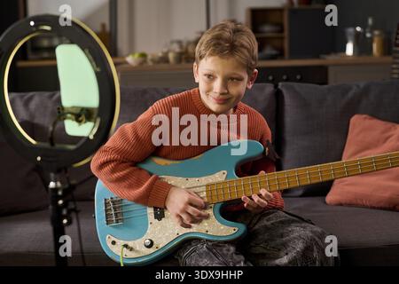 Caucasian boy playing electric bass guitar while sitting on sofa, smiling and looking slightly away from camera, recording music video using smartphone mounted on ring light Stock Photo