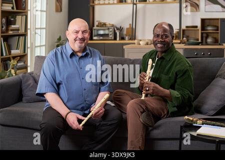 Middle aged Caucasian man and middle aged Black man sitting on sofa holding wooden recorders, smiling and looking toward camera, musical instruments resting on table nearby Stock Photo