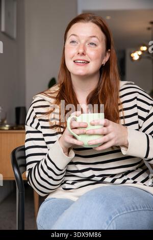 Fair-skinned red-haired woman sitting in armchair holding green mug and looking upward in lounge. Cozy, relaxation, warm, contemporary, lifestyle, comfort, domestic || Model released Stock Photo