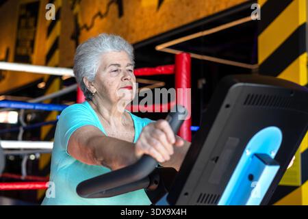 Active senior woman exercising on Stationary Bike in gym. Concept of menopause fitness, bone health, osteoporosis prevention and healthy aging. Stock Photo