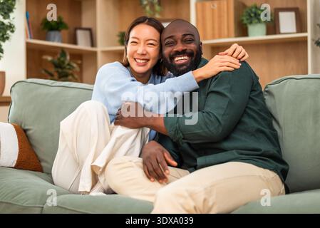 A joyful multiracial couple sits closely together on a green sofa, laughing and embracing in a cozy, modern living room. Stock Photo