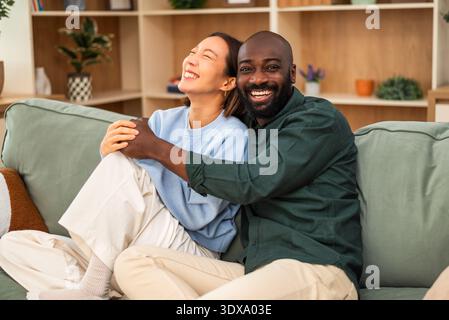 A happy interracial couple shares a candid moment of laughter while relaxing at home. Stock Photo