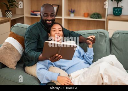 Happy multiracial couple embraces affectionately on a sofa, reading book and relaxing at home. Stock Photo