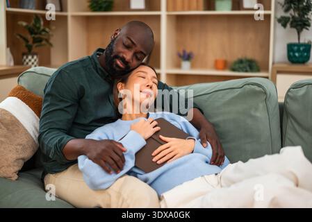 Multiracial couple embraces affectionately on a sofa, reading book and relaxing at home. Stock Photo