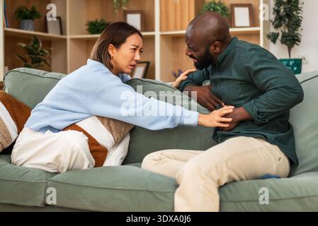 Adult multiracial couple sitting on sofa at home at talking about their health problems. Man is having stomachache. Stock Photo