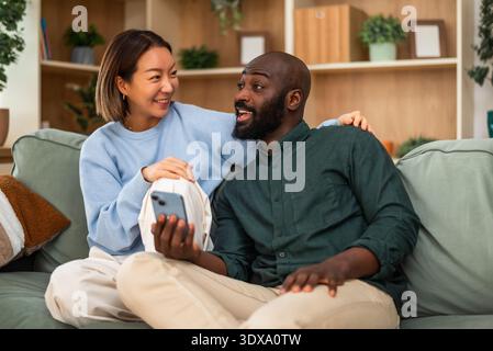 Happy and loving multiracial couple shares a joyful moment while relaxing at home. They are using smartphone. Stock Photo