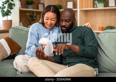 Happy and loving multiracial couple shares a joyful moment while relaxing at home. They are using smartphone. Stock Photo
