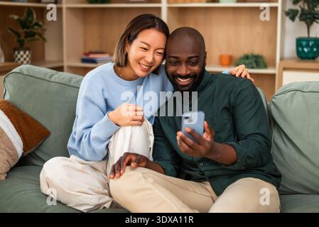 Happy and loving multiracial couple shares a joyful moment while relaxing at home. They are using smartphone. Stock Photo