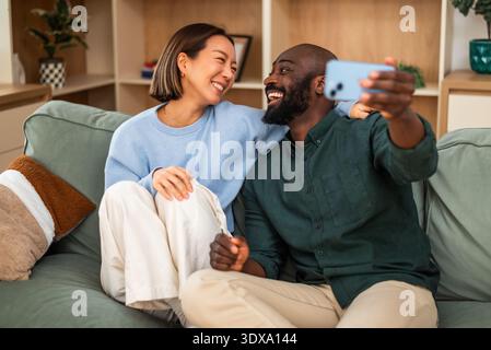 A joyful multiracial couple sits closely together on a green sofa, laughing and embracing in a cozy, modern living room. They are taking selfie. Stock Photo