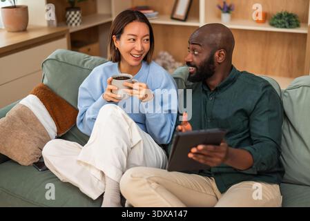 Happy interracial couple shares a joyful moment while relaxing at home. They are drinking coffee,talking and using digital tablet. Stock Photo