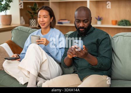 Multiracial couple relaxing at home. Woman is watching tv and man is using smartphone. Stock Photo
