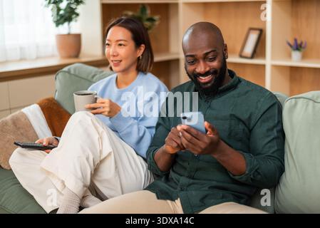 Happy multiracial couple shares a joyful moment while relaxing at home. Woman is watching tv and man is using smartphone. Stock Photo