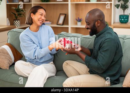 Cheerful multiracial couple is having fun at home while while exchanging gifts. Stock Photo