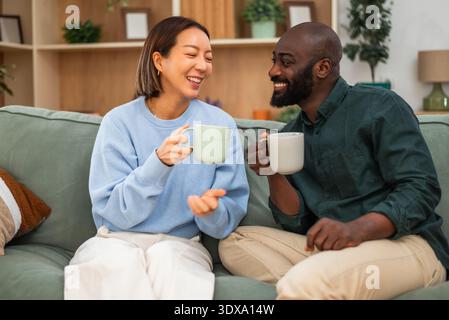 Happy interracial couple shares a joyful moment while relaxing at home. They are drinking coffee and talking. Stock Photo