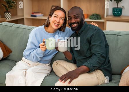 Happy interracial couple shares a joyful moment while relaxing at home. They are drinking coffee and looking at camera. Stock Photo