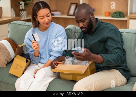 Multiracial couple is sitting on couch at home and shopping online. Man is holding smartphone and wife is holding payment card. Stock Photo