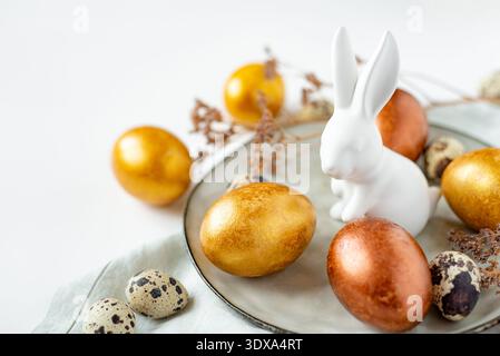 Golden and bronze Easter eggs and Easter bunny in a gray plate on a linen napkin on a white background Stock Photo