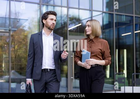 Two professional business colleagues, a man and a woman, are walking and engaging in conversation outside a modern corporate office building, discussing work Stock Photo