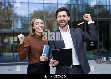 Two excited business colleagues celebrating a professional victory, raising their fists in triumph while standing outside a modern office building, representing teamwork and accomplishment Stock Photo