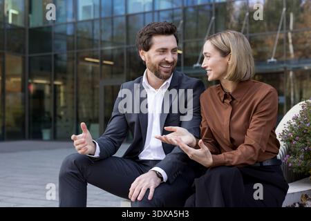 Two smiling business professionals engaging in a lively outdoor conversation, discussing concepts and networking while sitting against a modern office building Stock Photo