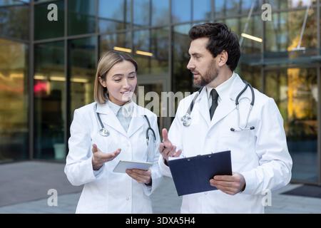 Two doctors confer outside a modern clinic, reviewing a tablet and clipboard as they discuss patient care, diagnosis and treatment plans in a professional teamwork setting Stock Photo