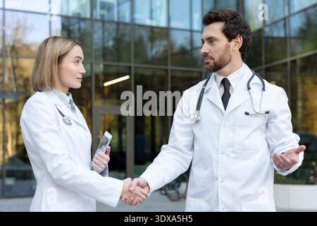 Doctors in white coats and stethoscopes shaking hands outside a modern clinic building, symbolizing a strong professional partnership, collaboration, and trust in healthcare and medicine Stock Photo