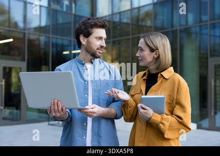 Startup team members having a productive business discussion and sharing ideas about a project while standing outside a modern office building, using a laptop and a tablet Stock Photo