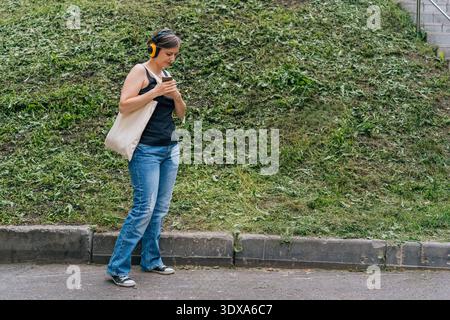 Confident blonde woman 40s in headphones carrying tote bag walking in city park Stock Photo