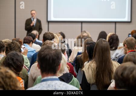 Crowd of attendees listening to speaker during business conference presentation, focused audience, seminar networking, learning atmosphere. Stock Photo
